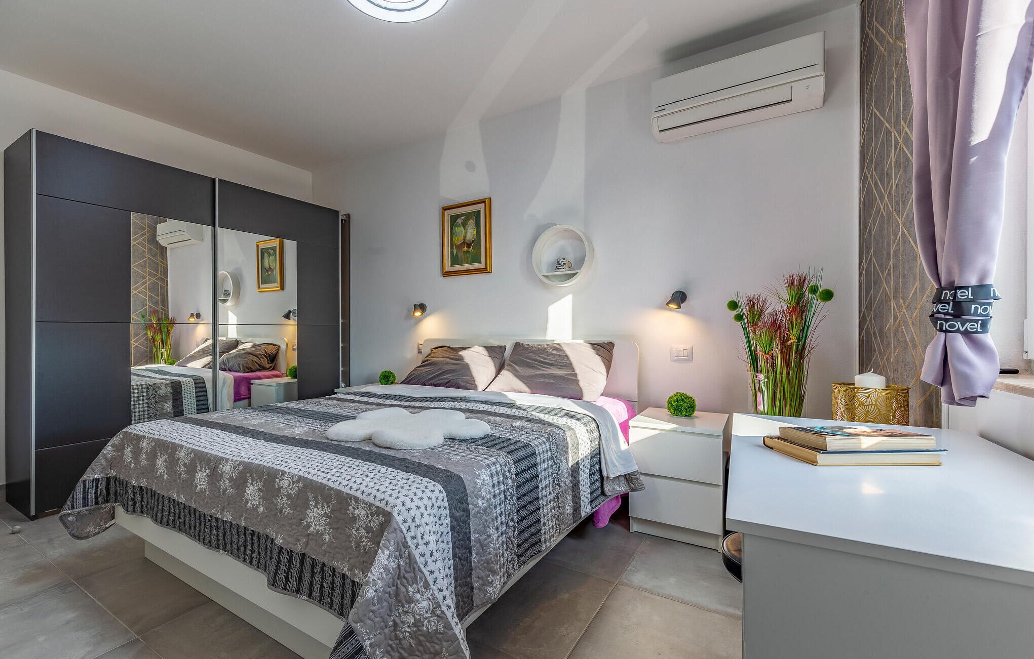 Un dormitorio moderno con una cama grande, mesitas de noche, un aparador, un espejo, un equipo de aire acondicionado y un jarrón con flores.
