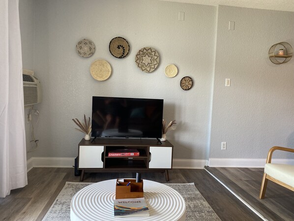 Una sala de estar con un televisor de pantalla plana, un sofá blanco, una mesa de centro de madera con una tarjeta de bienvenida y decoración en la pared.