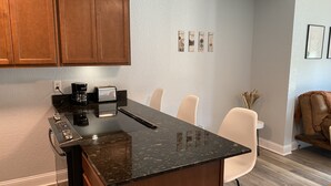 Una cocina con encimeras oscuras, armarios de madera, cafetera y tostadora. Hay cuatro sillas y un jarrón con plantas secas en la pared.