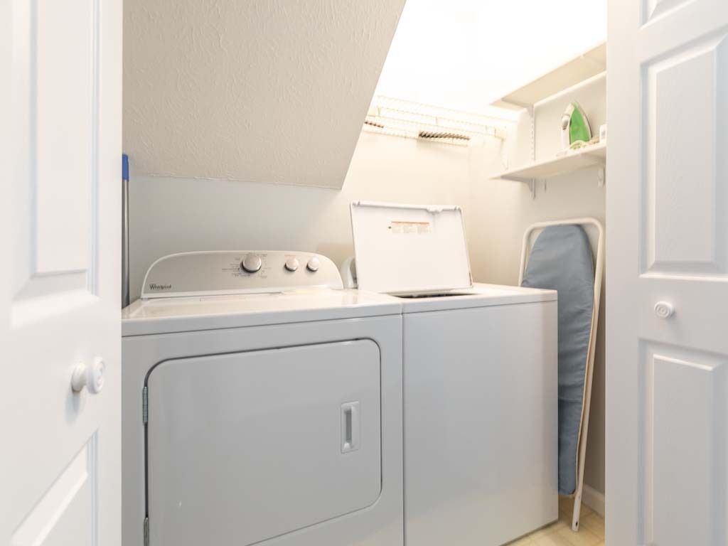 A compact laundry area with a front-loading washer and dryer, a small shelf with a green item, and a door leading to another room.