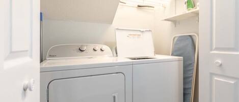 A compact laundry area with a front-loading washer and dryer, a small shelf with a green item, and a door leading to another room.