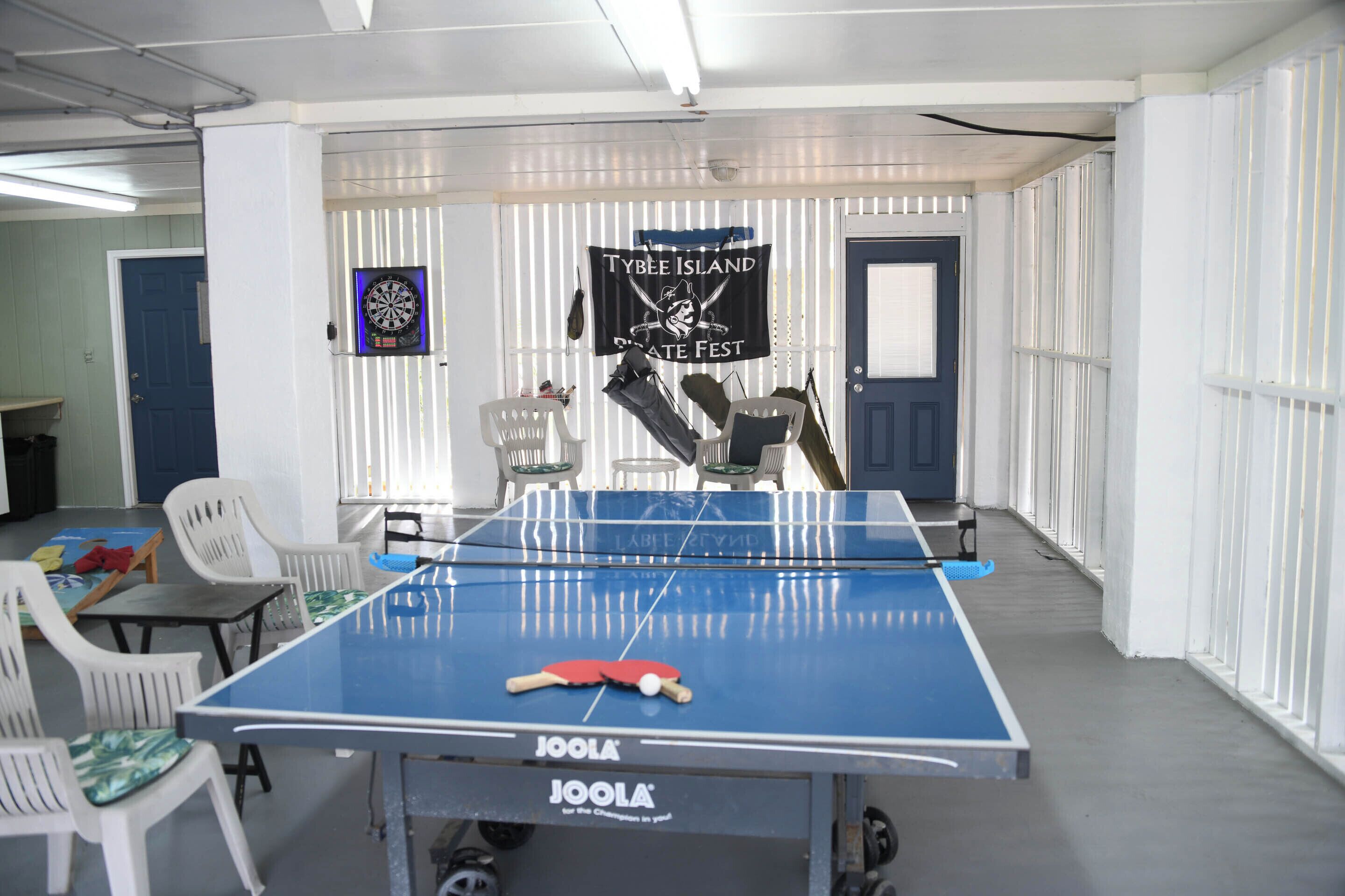 Una habitación con mesa de ping pong, sillas de plástico blanco y dardo.