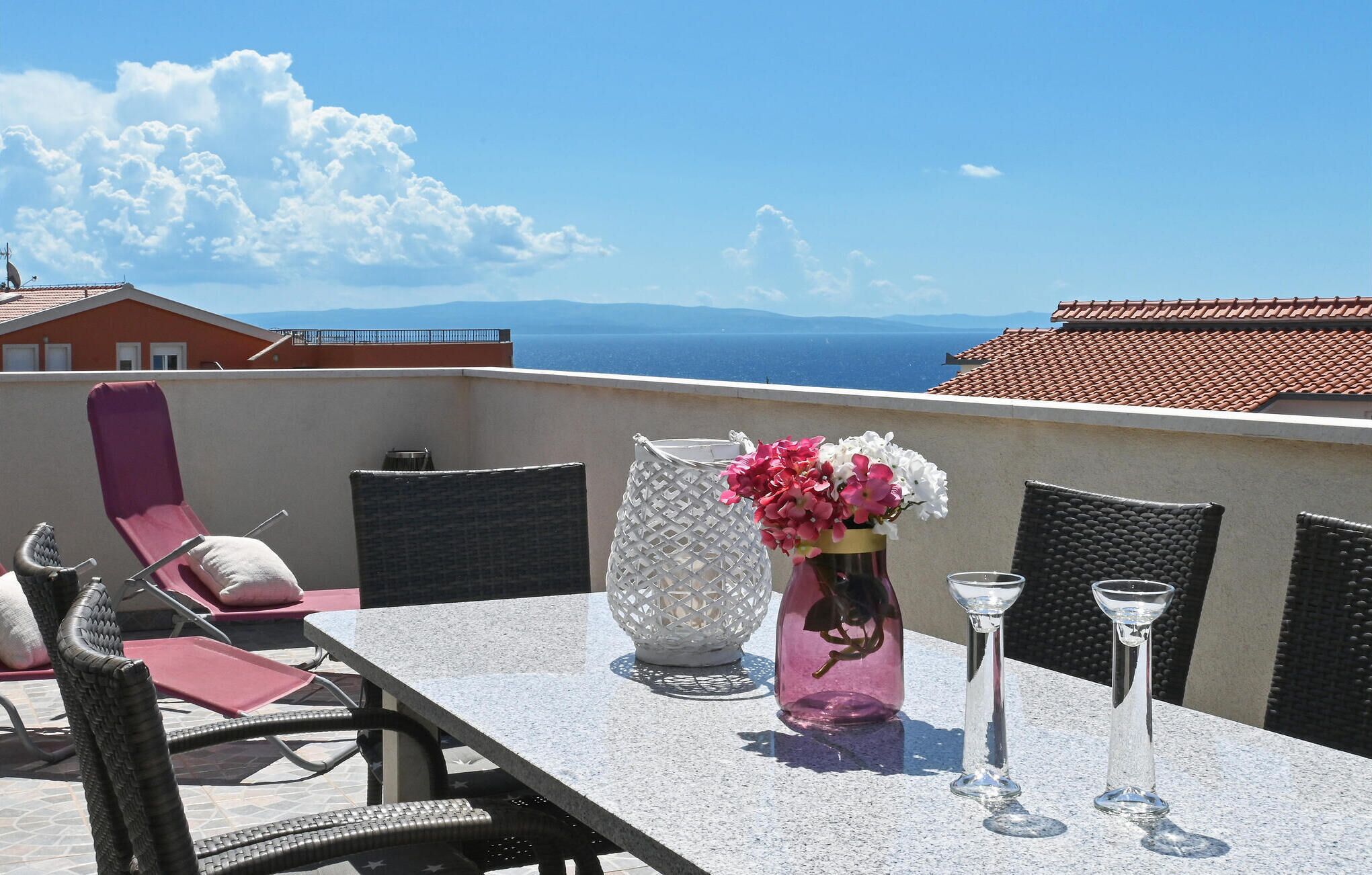 A balcony with a table, chairs, a vase with flowers, and a view of the sea and mountains.