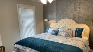 A bedroom with a tufted headboard, a large bed with a teal quilt and floral bedding, a wooden chair, a window with blinds, and a textured wall. - Dubuque Luxury Rentals Washington Street (Dubuque)