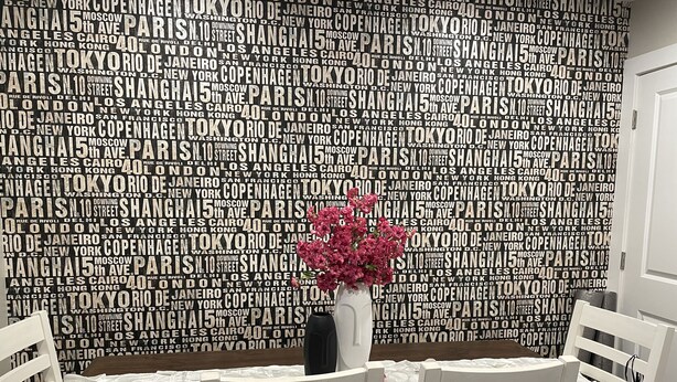 A dining area with a table set for two, surrounded by white chairs. A vase with pink flowers is placed on the table. The wall behind is covered with a pattern of city names in a repeating design. - Dubuque Luxury Rentals Washington Street (Dubuque)