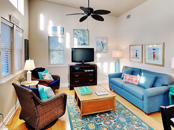 A living room with a television, blue sofa, wicker chair, and a ceiling fan. - Treetop Cottage by Tybee Vacation Rentals (Tybee Island)