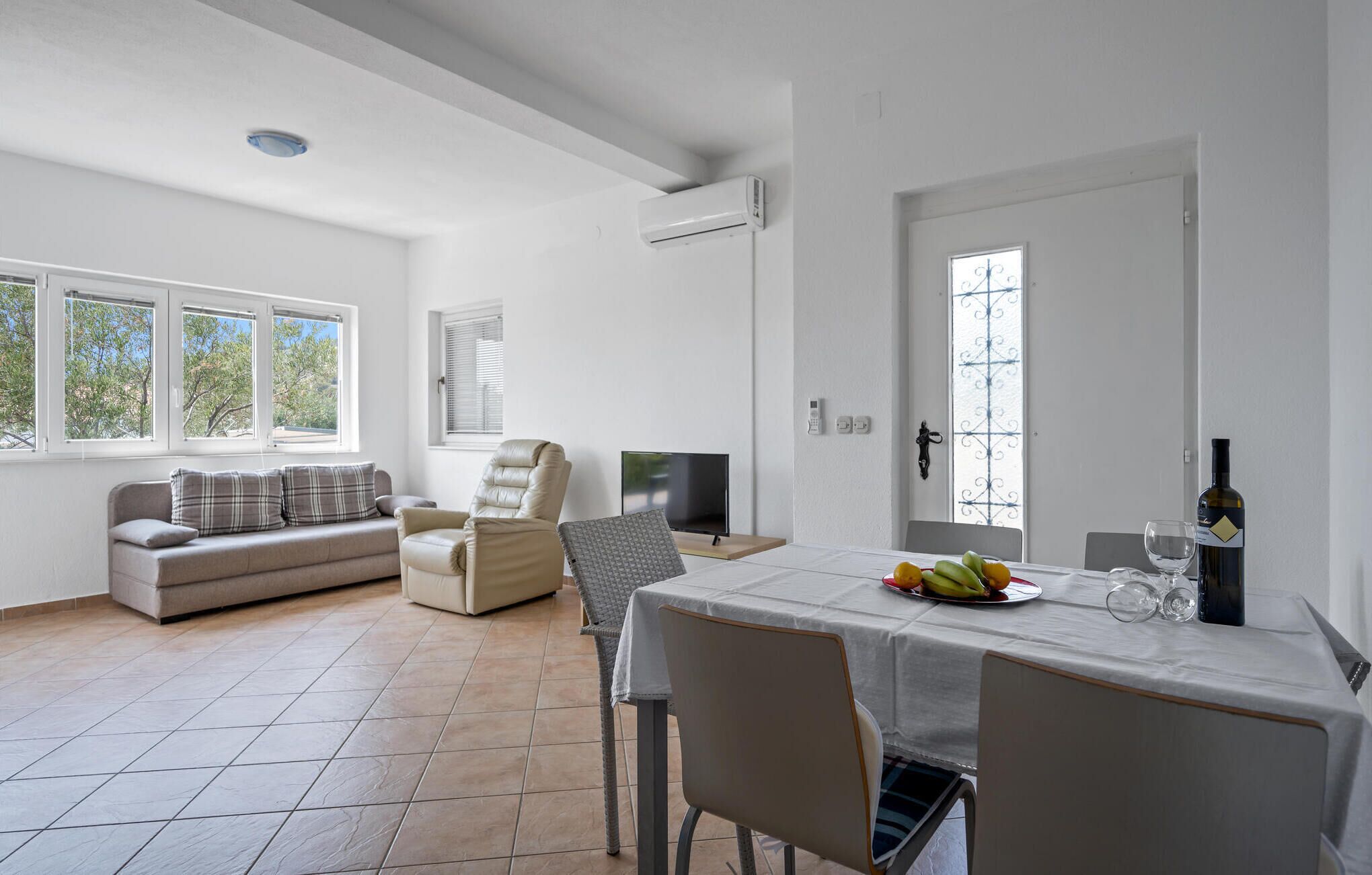 En rummelig opholdsstue med sofa, spisebord dækket med frugt og en flaske samt et fjernsyn.