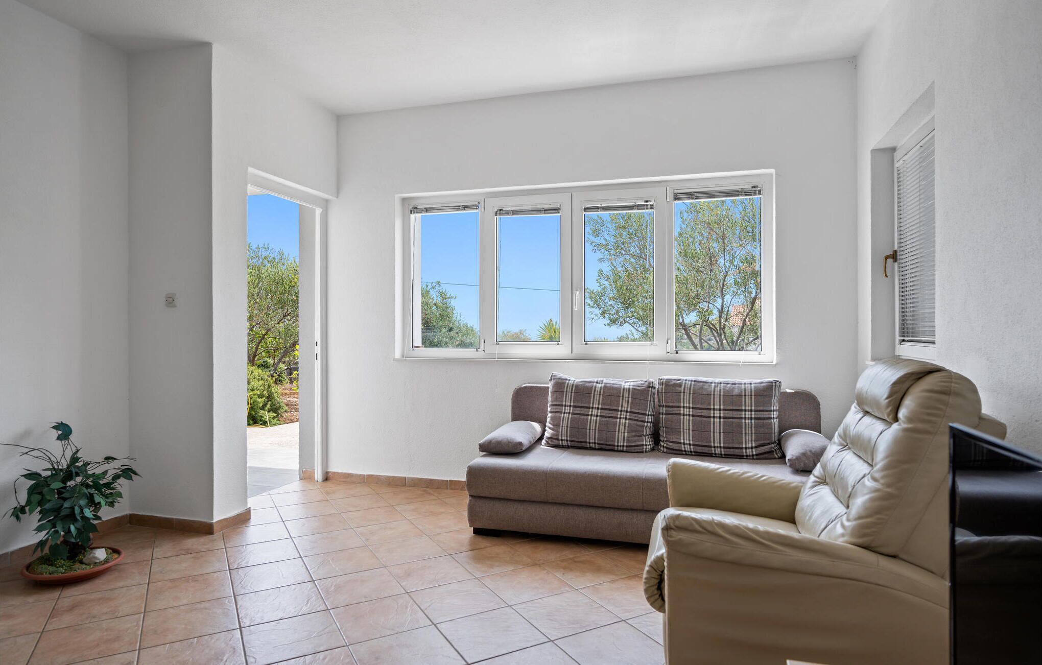 A spacious living room with a beige sofa, a small potted plant, and a window with a view of trees and a clear sky.