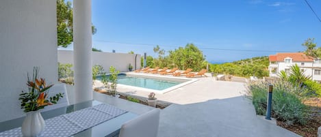 A modern outdoor pool area with lounge chairs, a glass table, and a view of the sea.
