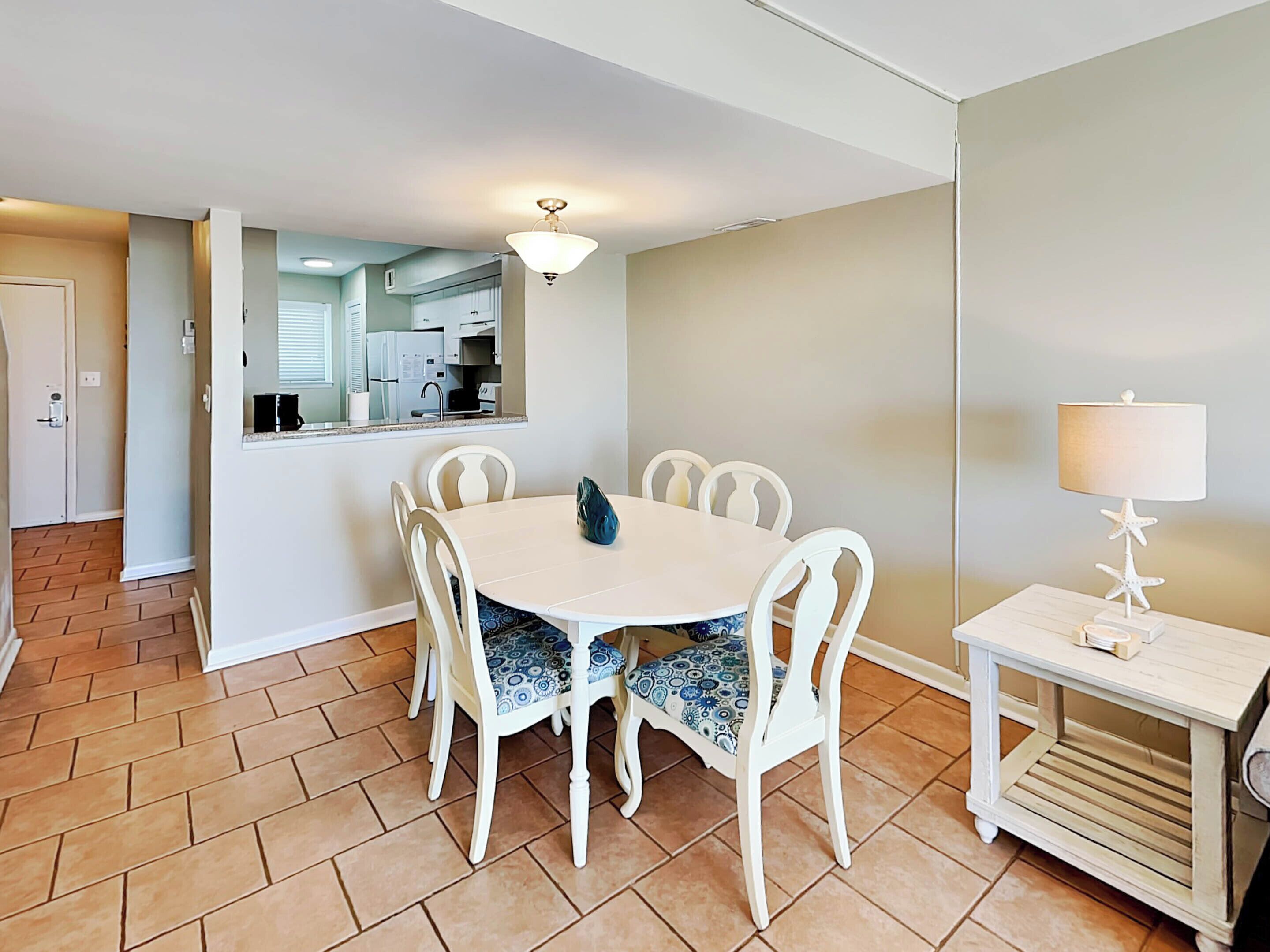 A dining area with a round table and white chairs, a small side table with a lamp, and a kitchen visible through an open door.