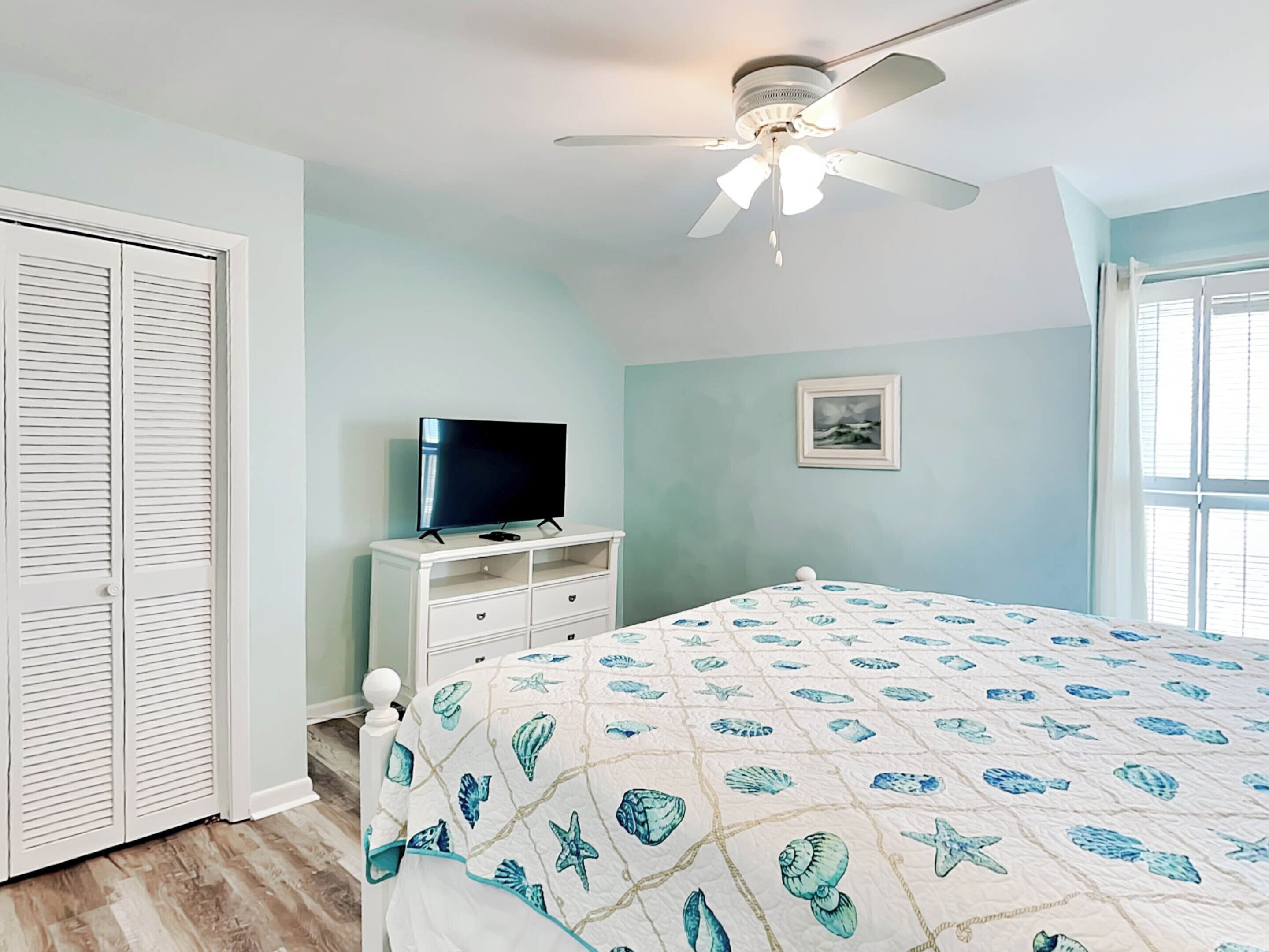 Un dormitorio con cama, ventilador de techo, televisor en una cómoda y ventana.