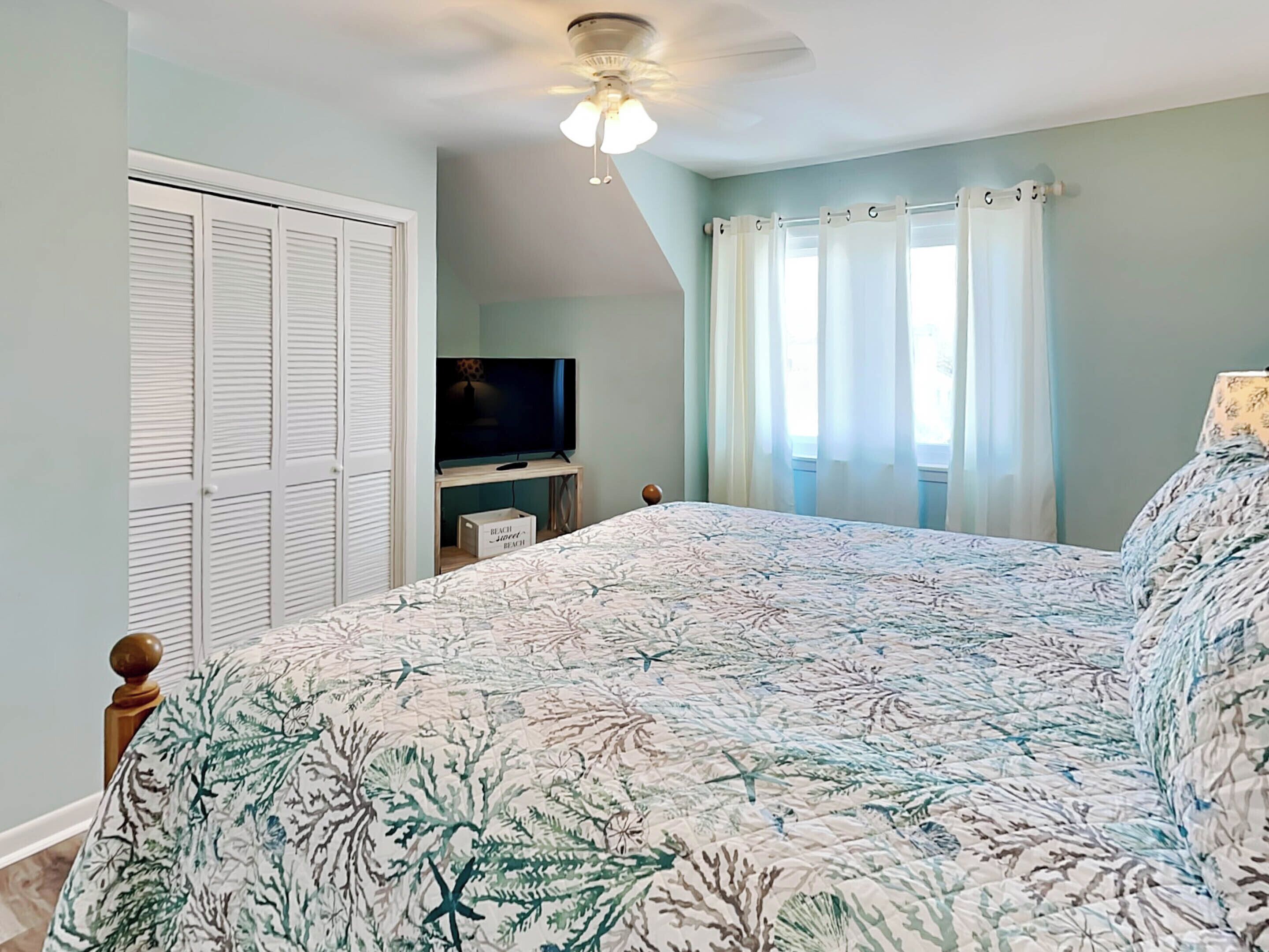 A bedroom with a bed, a ceiling fan, a television on a stand, and a window with curtains.