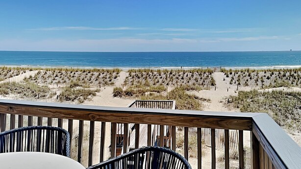 A beach view from a wooden deck with chairs, featuring a sandy beach and ocean. - Tybee Lights 112B by Tybee Vacation Rentals (Tybee Island)