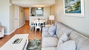 A living room with a grey sofa, a white coffee table, and a dining area with a table and chairs. - Tybee Lights 112B by Tybee Vacation Rentals (Tybee Island)
