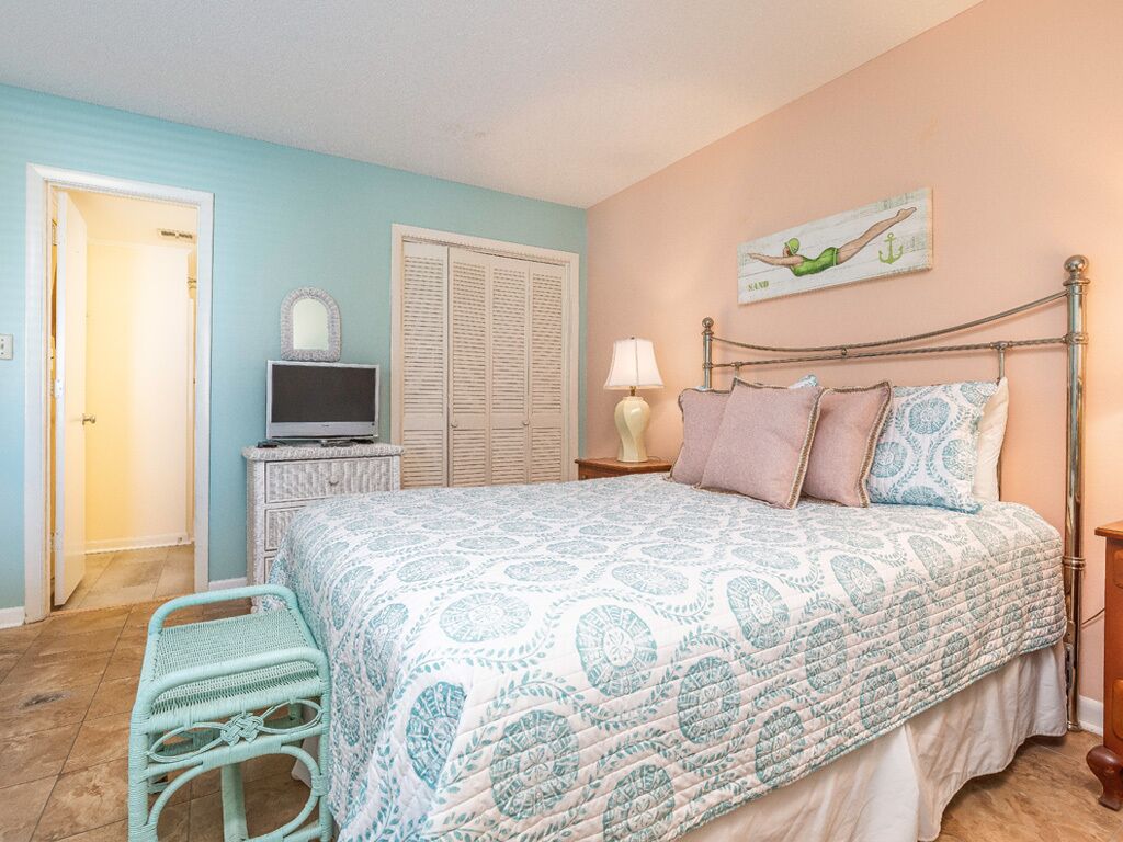 A bedroom with a bed, a nightstand, a lamp, a chair, a dresser, a television, and a door.