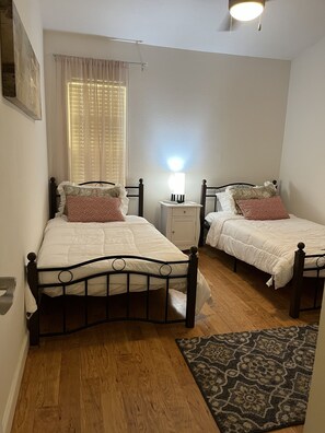 Een slaapkamer met twee eenpersoonsbedden, een nachtkastje, een bedlampje, een raam met luxaflex en een kamerbreed tapijt met een patroon.
