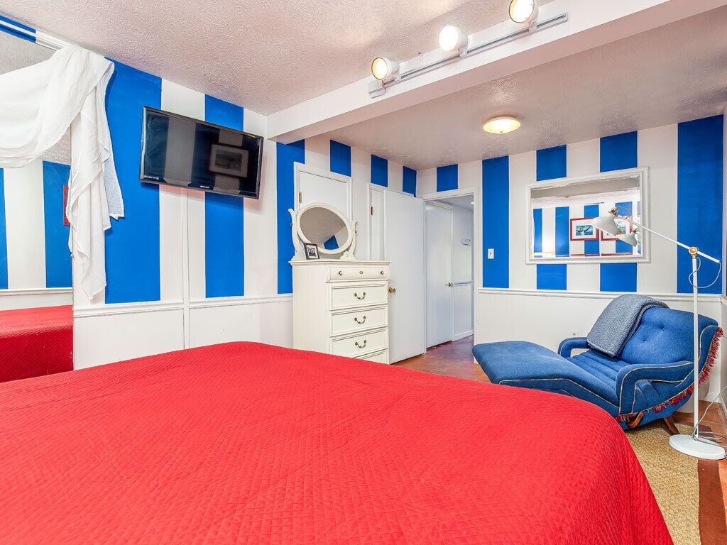 Een kamer met een rode bed, een blauwe fauteuil, een witte kaptafel, een spiegel, een televisie en een raam met jaloezieën.