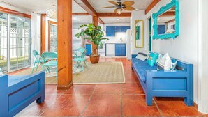A spacious living area with a blue sofa, a blue bench, and a ceiling fan. There is a large window providing natural light and a view of the outside. - Tybreeza A by Tybee Vacation Rentals (Tybee Island)