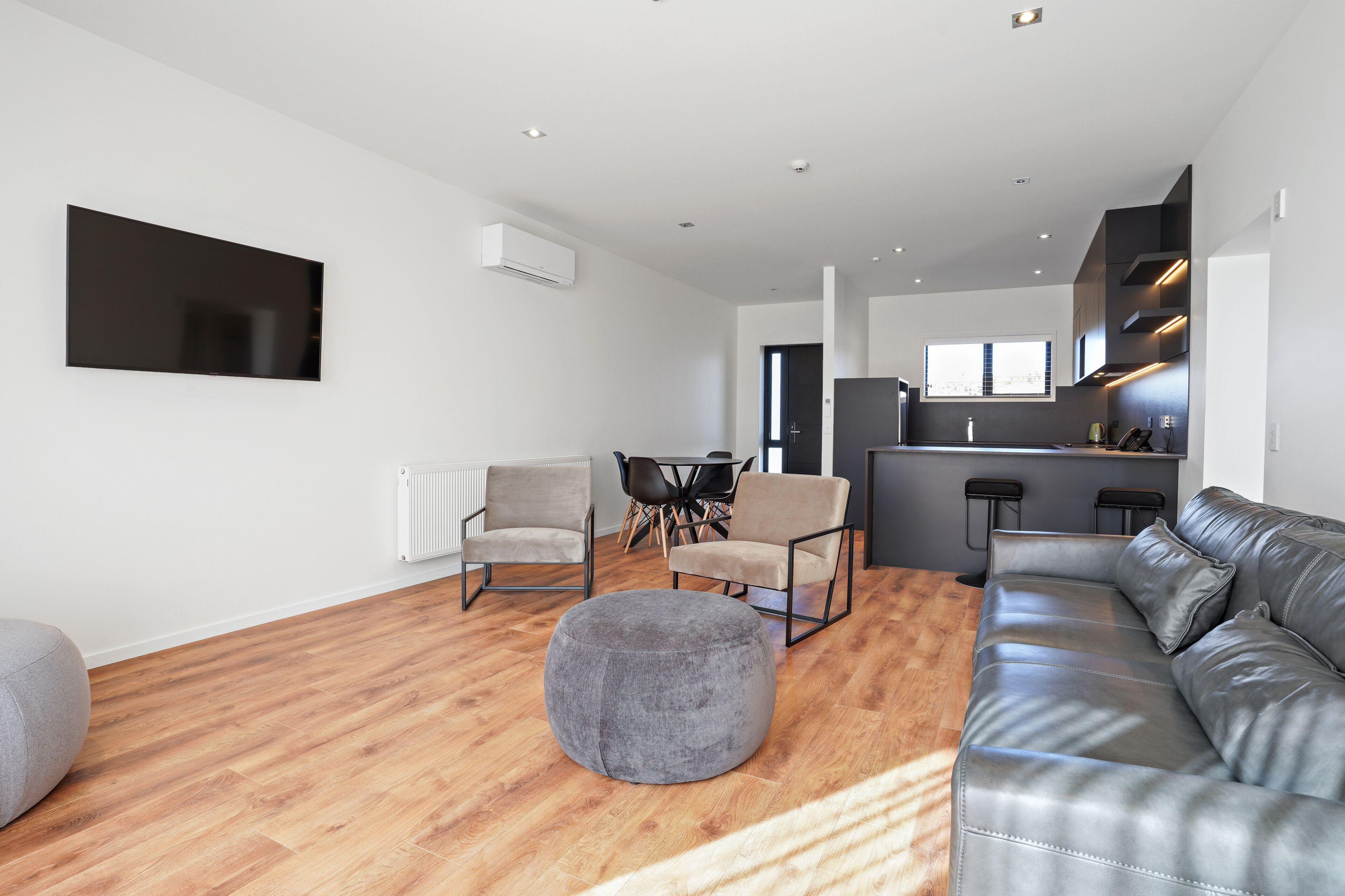 A modern living room with a grey sofa, a grey ottoman, and a wooden floor. There is a dining area with a table and chairs, and a kitchen area with a bar counter.