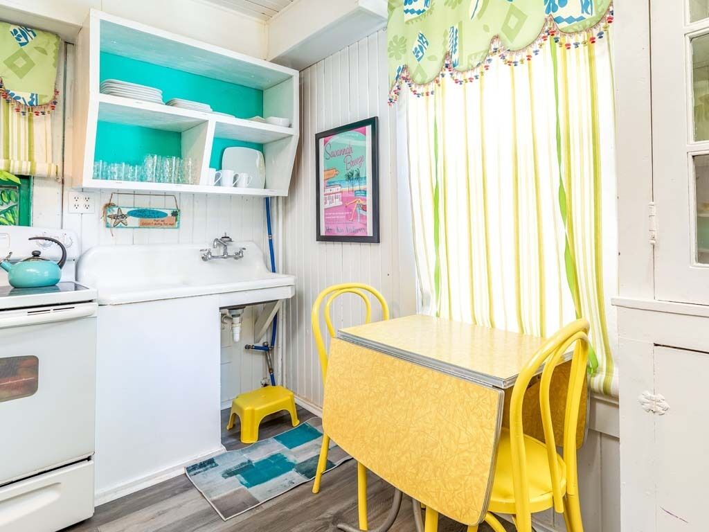 Cocina pequeña con armarios blancos, un lavamanos y un taburete. Un comedor amarillo con sillas está ubicado cerca de una ventana con cortinas a rayas.