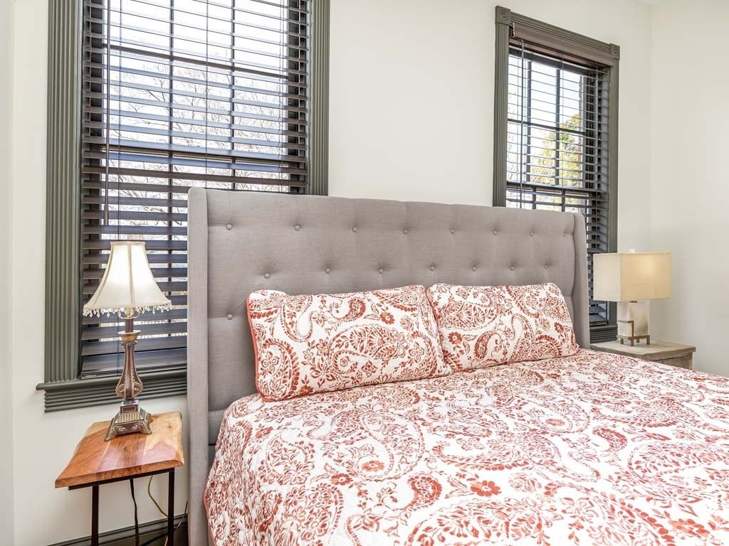 A bedroom with a tufted headboard, patterned bedding, two bedside lamps, and windows with blinds.