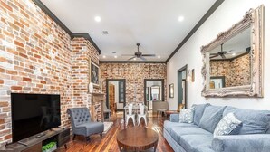 A living room with a brick wall, a flat-screen TV, a grey sofa, a round coffee table, and a fireplace. - Waldburg Terrace by Tybee Vacation Rentals (Savannah)