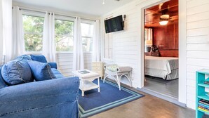 Une pièce avec un canapé bleu, une petite table, une chaise en osier, une bibliothèque et un téléviseur fixé au mur.