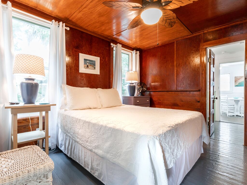 A bedroom with a large bed, wooden walls, a ceiling fan, and a window with curtains.