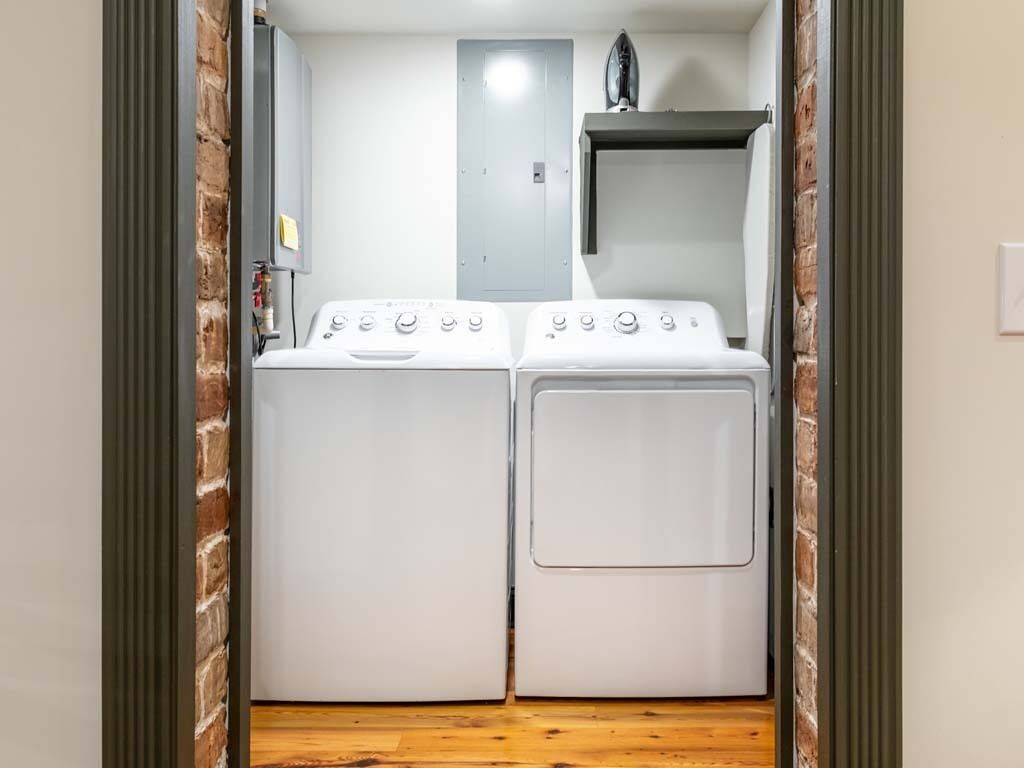A laundry room with two white washing machines and a dryer, a brick wall, and a door leading to another room.
