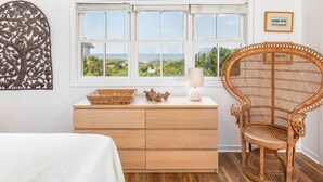 Un dormitorio con cómoda de madera, una silla de mimbre, una ventana con vistas y un adorno en la pared.