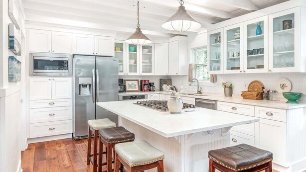 A kitchen with white cabinets, a stainless steel refrigerator, a large island with stools, and pendant lighting. - Wrens Nest by Tybee Vacation Rentals (Tybee Island)