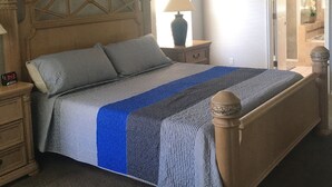 Un dormitorio con cabecera de madera, una cama con manta azul y gris a rayas, una mesita de noche con lámpara y un ventilador de techo.