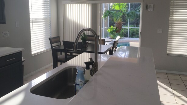Una cocina con una isla grande, un comedor con mesa y sillas, y vista a una piscina y palmeras a través de las ventanas.