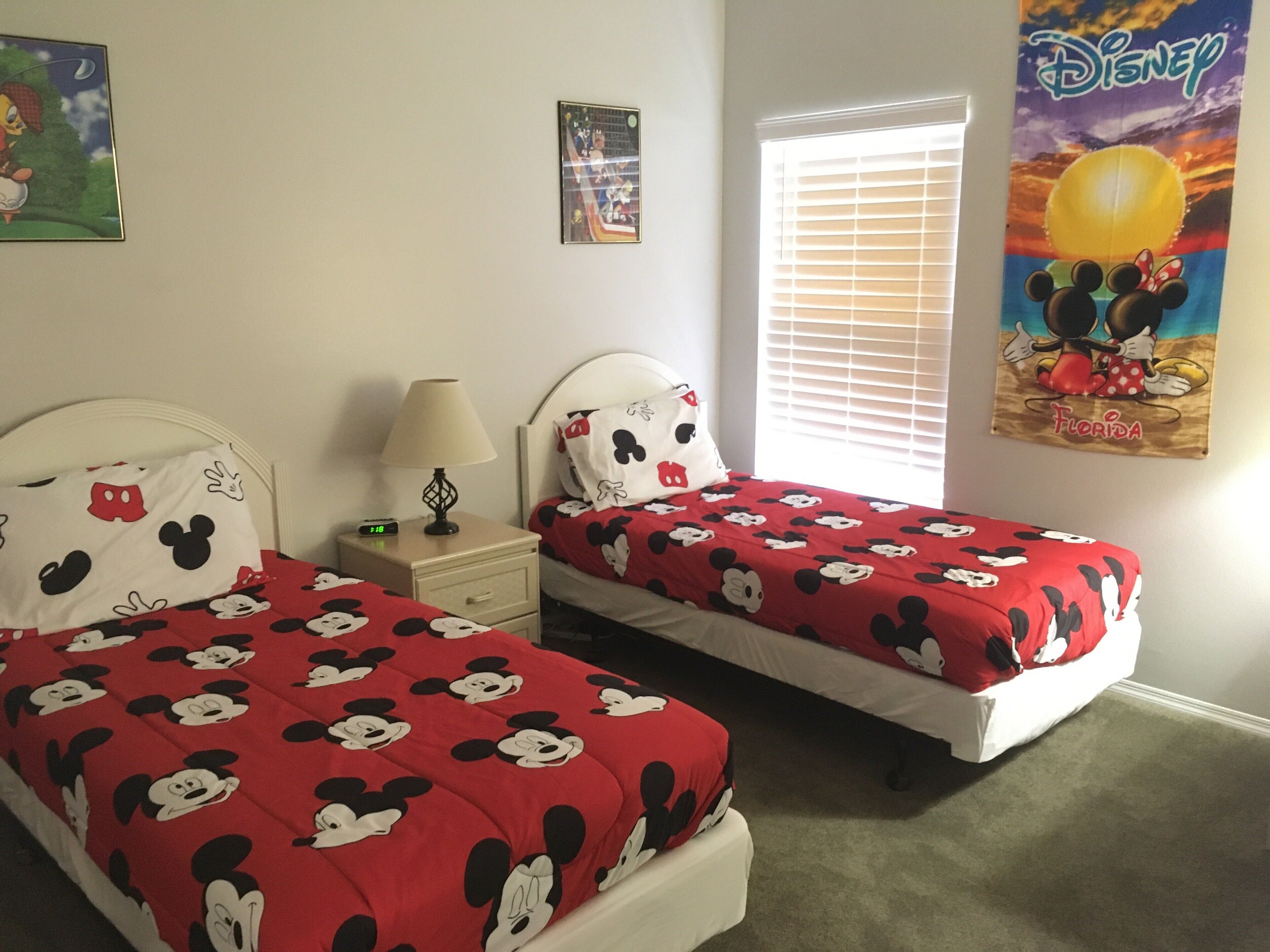 Une chambre d’hôtel avec deux lits, chacun agrémenté de draps et de couvertures aux motifs de Mickey Mouse. On y trouve une table de chevet avec une lampe et une horloge, ainsi qu’une affiche de Mickey Mouse accrochée au mur.