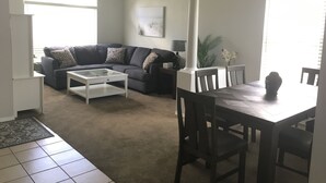 Una sala de estar con un sofá de color oscuro, una mesa de centro blanca, una mesa de comedor con sillas, un pilar blanco y una ventana con persianas.