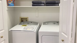 A laundry room with two front-loading washing machines, a shelf with folded towels, and a Tide detergent box.