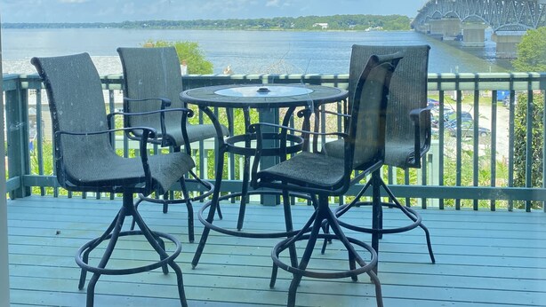 A covered outdoor seating area with a table and chairs, overlooking a body of water and a bridge.