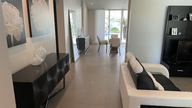 A modern living room with a white sofa, black and white armchairs, a black console table, and a large mirror. There is a dining area in the background with a table and chairs. - Luxurious modern 3-bedroom plus den home w/ pool & hot tub (Phoenix)