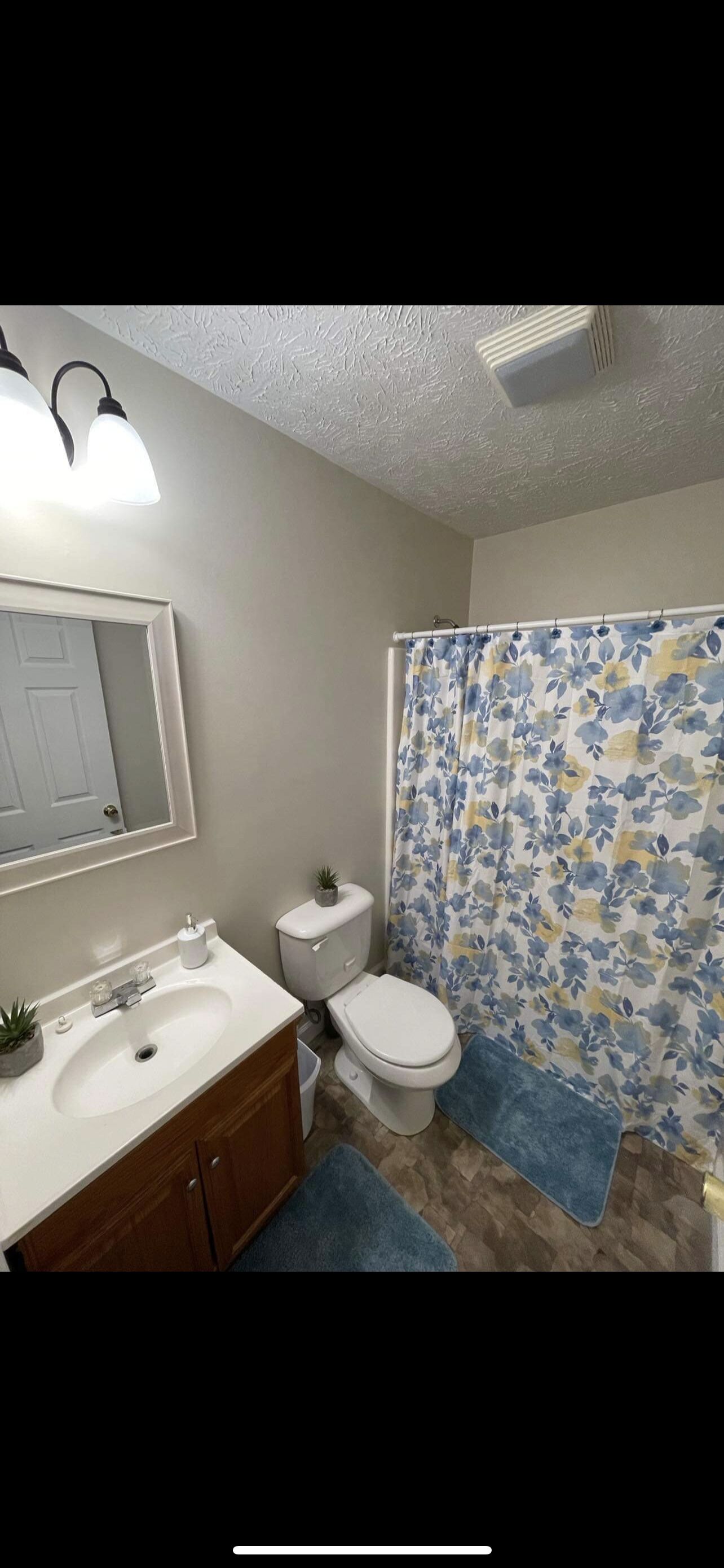Un baño con lavamanos blanco, espejo y inodoro. Hay una cortina de ducha con estampado floral.
