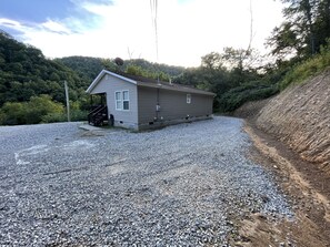 Simple, single-story building with a front porch and staircase, situated on a gravel driveway amidst a natural landscape.