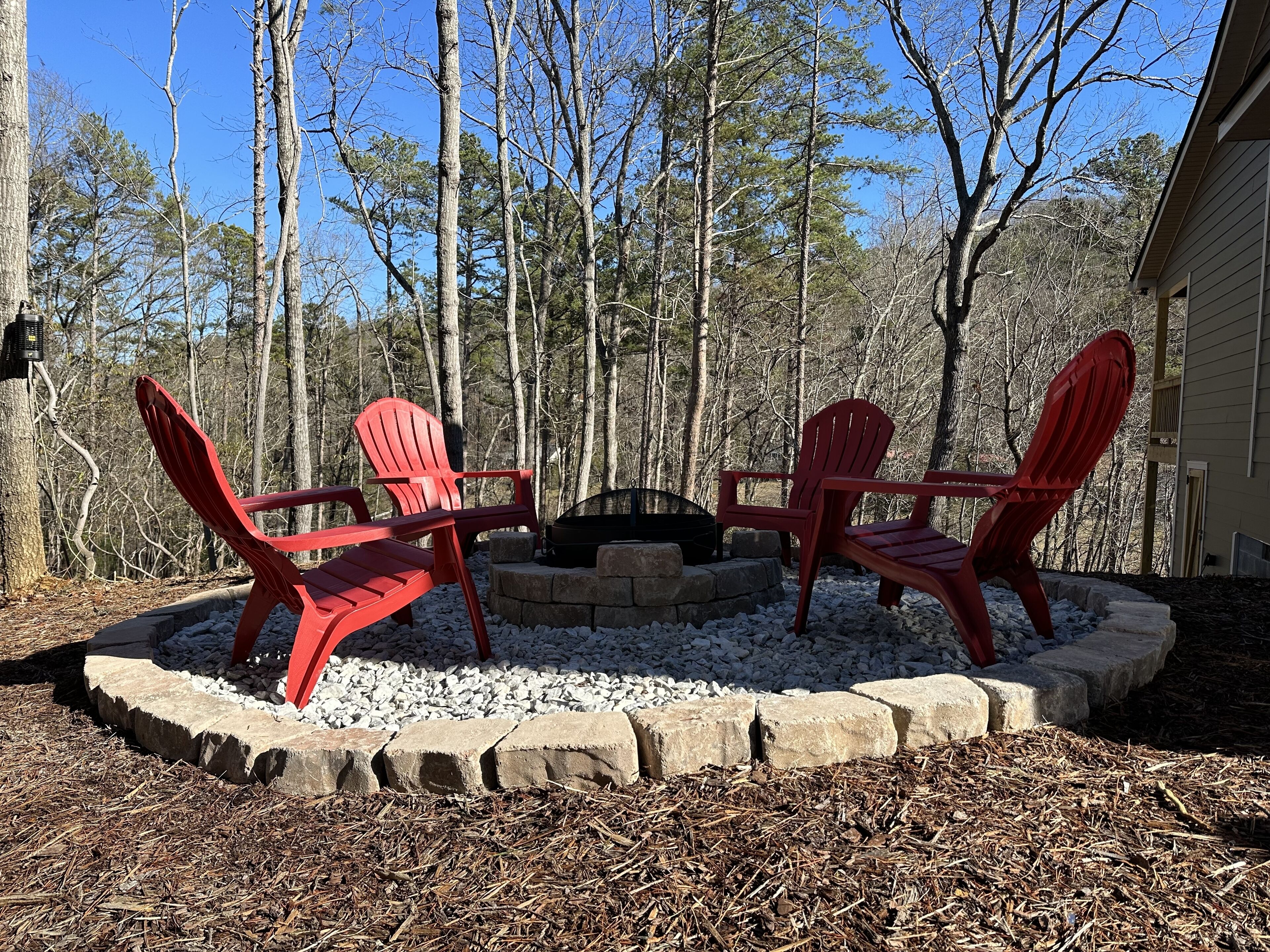 A circular fire pit with two red Adirondack chairs facing it, surrounded by a stone border, set in a wooded area with a house in the background.