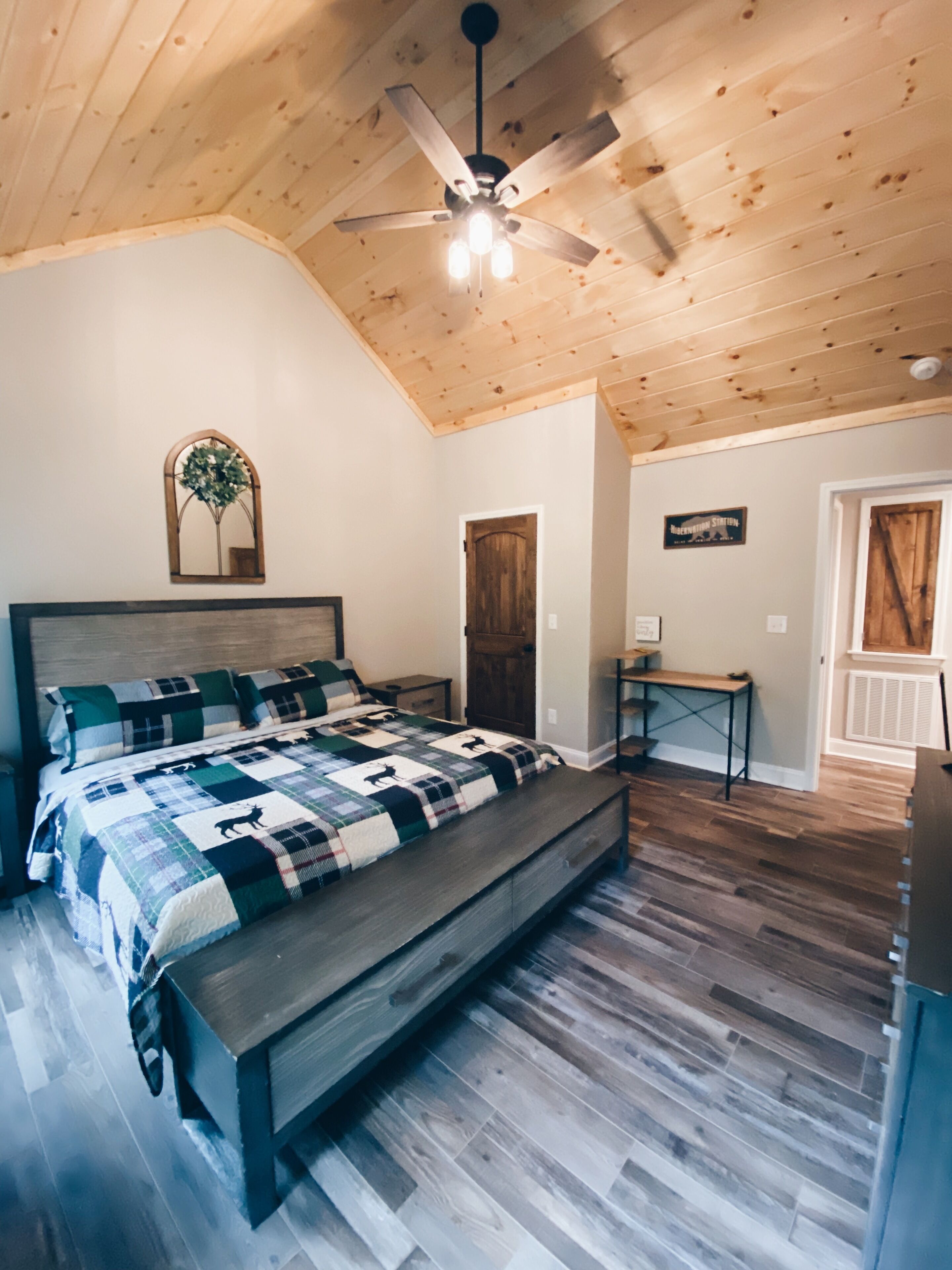 Une chambre avec un plafond en bois, un lit recouvert d’une couverture à carreaux, une petite table et une porte.