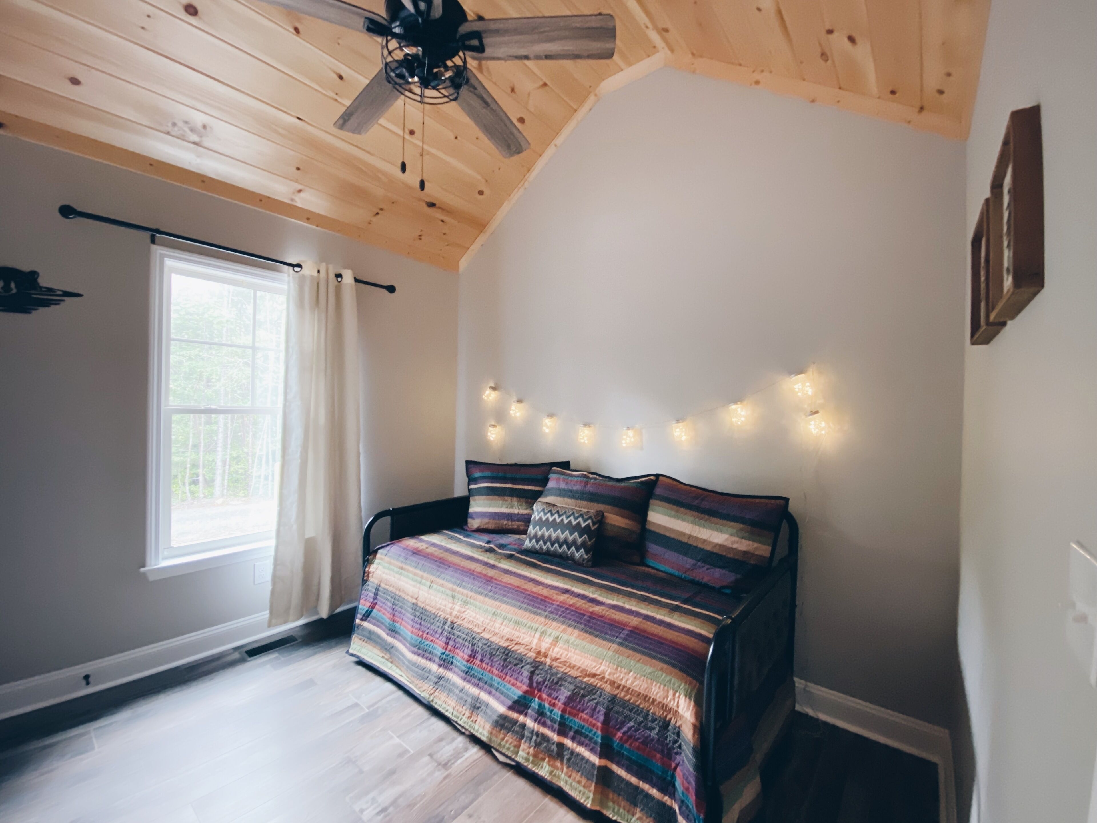 Una camera da letto con soffitto spiovente, un letto con copripiumino a righe colorate, una ventola a soffitto, una finestra con tende e luci a stringa sulla parete.