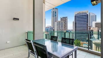 Un balcón con una mesa y sillas de vidrio, con vista a un paisaje urbano de edificios modernos.