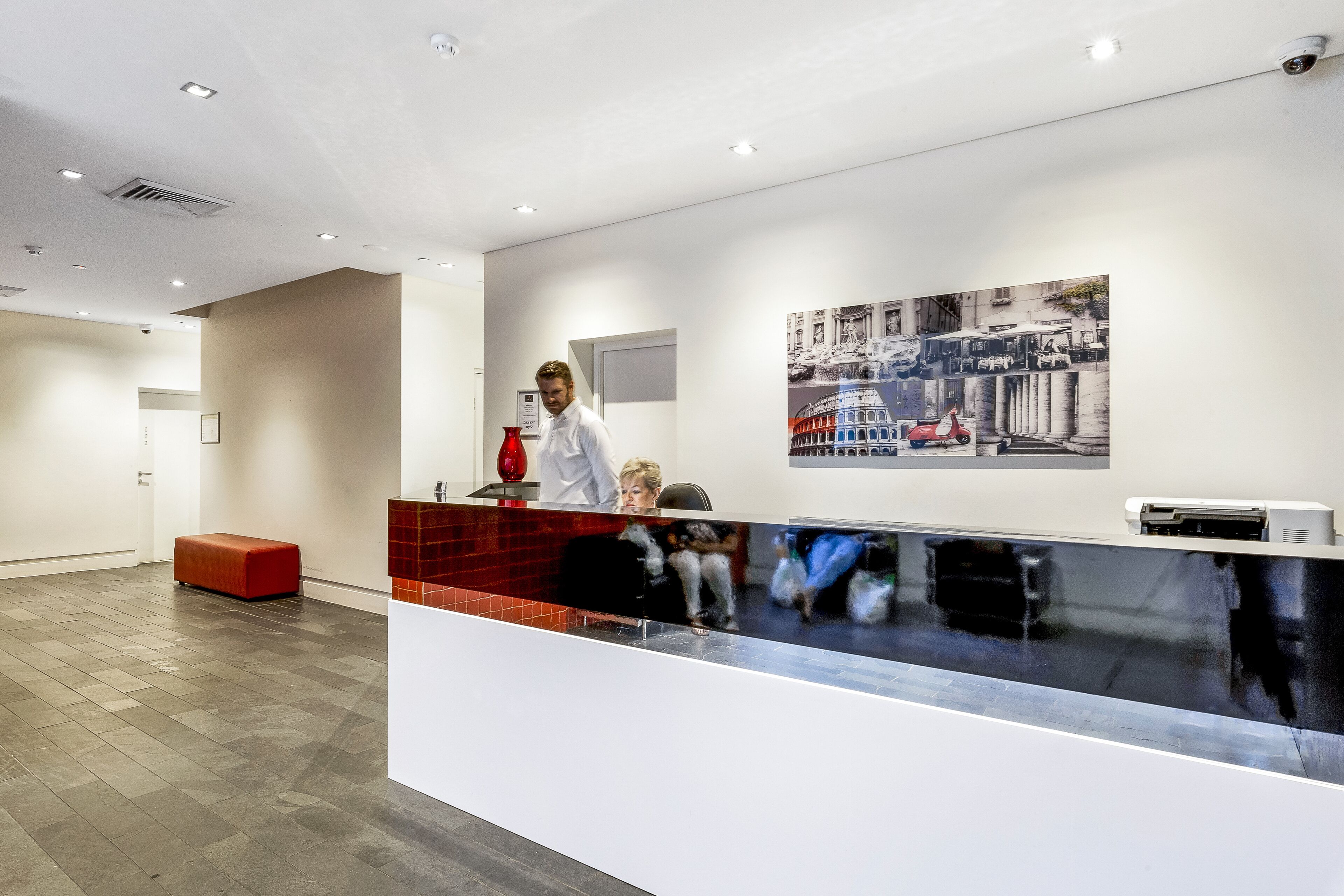Un área de recepción de diseño moderno, con mostrador de recepción, un banco rojo e impresora mural.