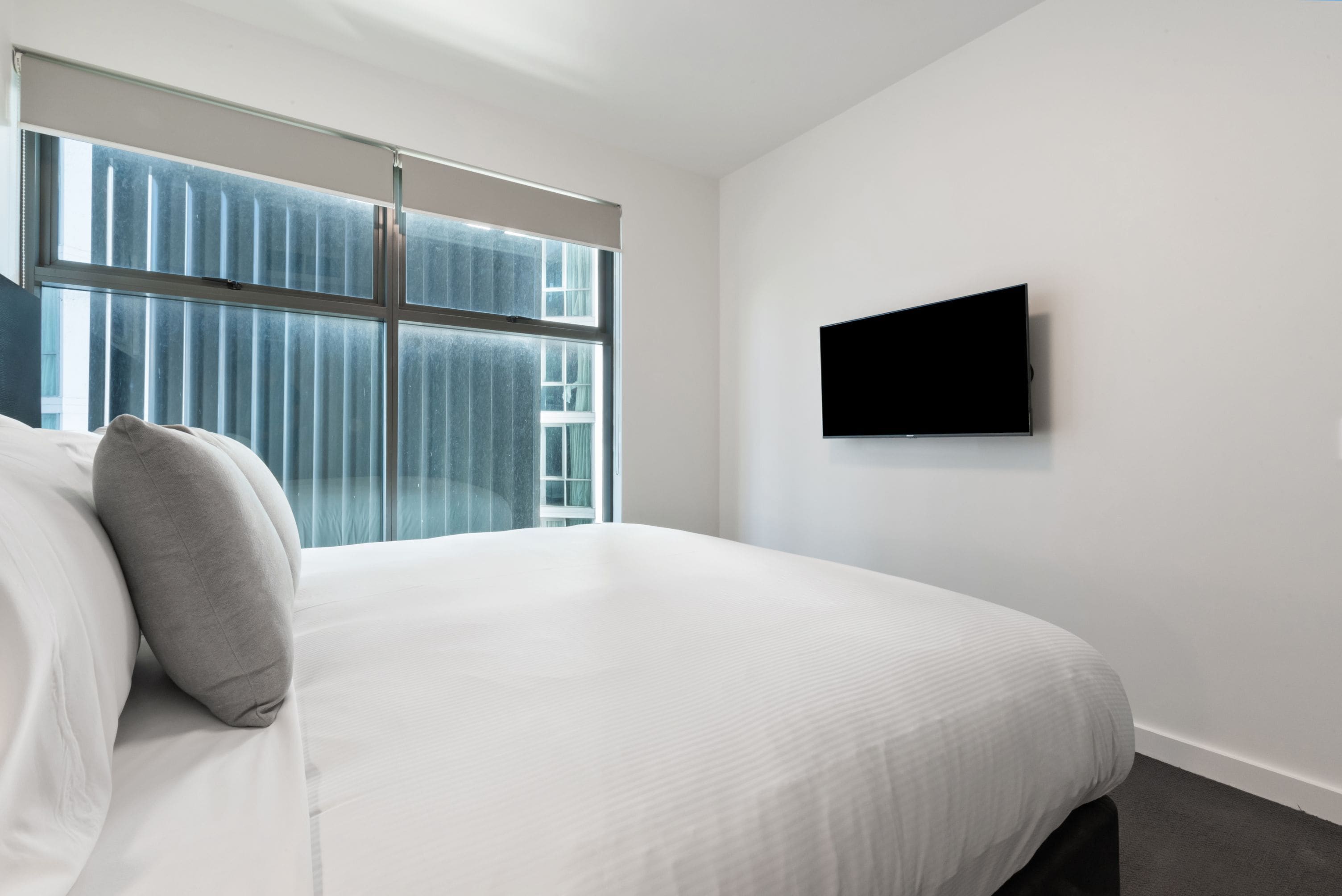 A modern hotel room with a large bed, a flat-screen TV mounted on the wall, and a large window with blinds.