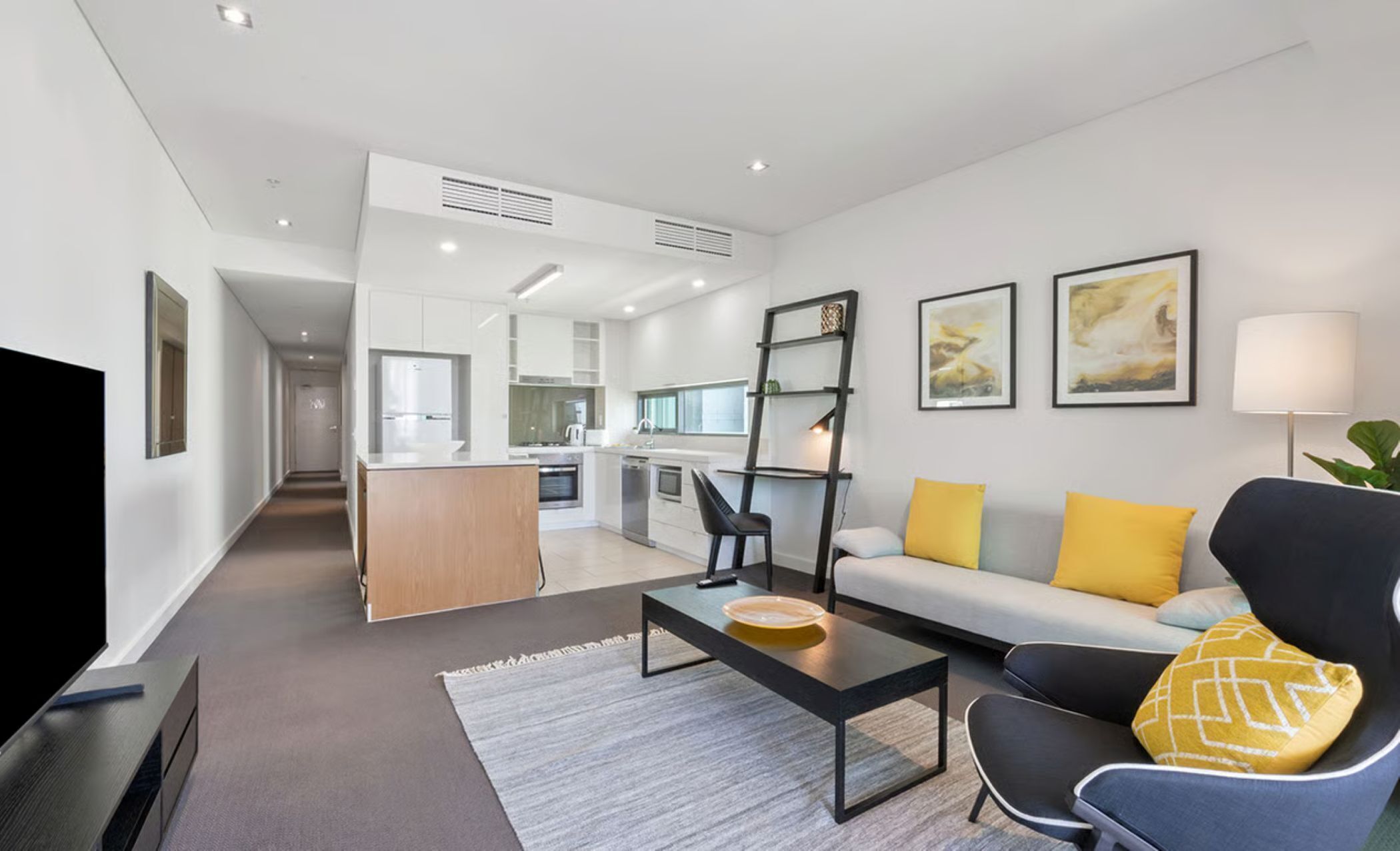 Una sala moderna con sofá, mesa de centro y televisor. Se aprecia una cocina al fondo con zona de comedor.