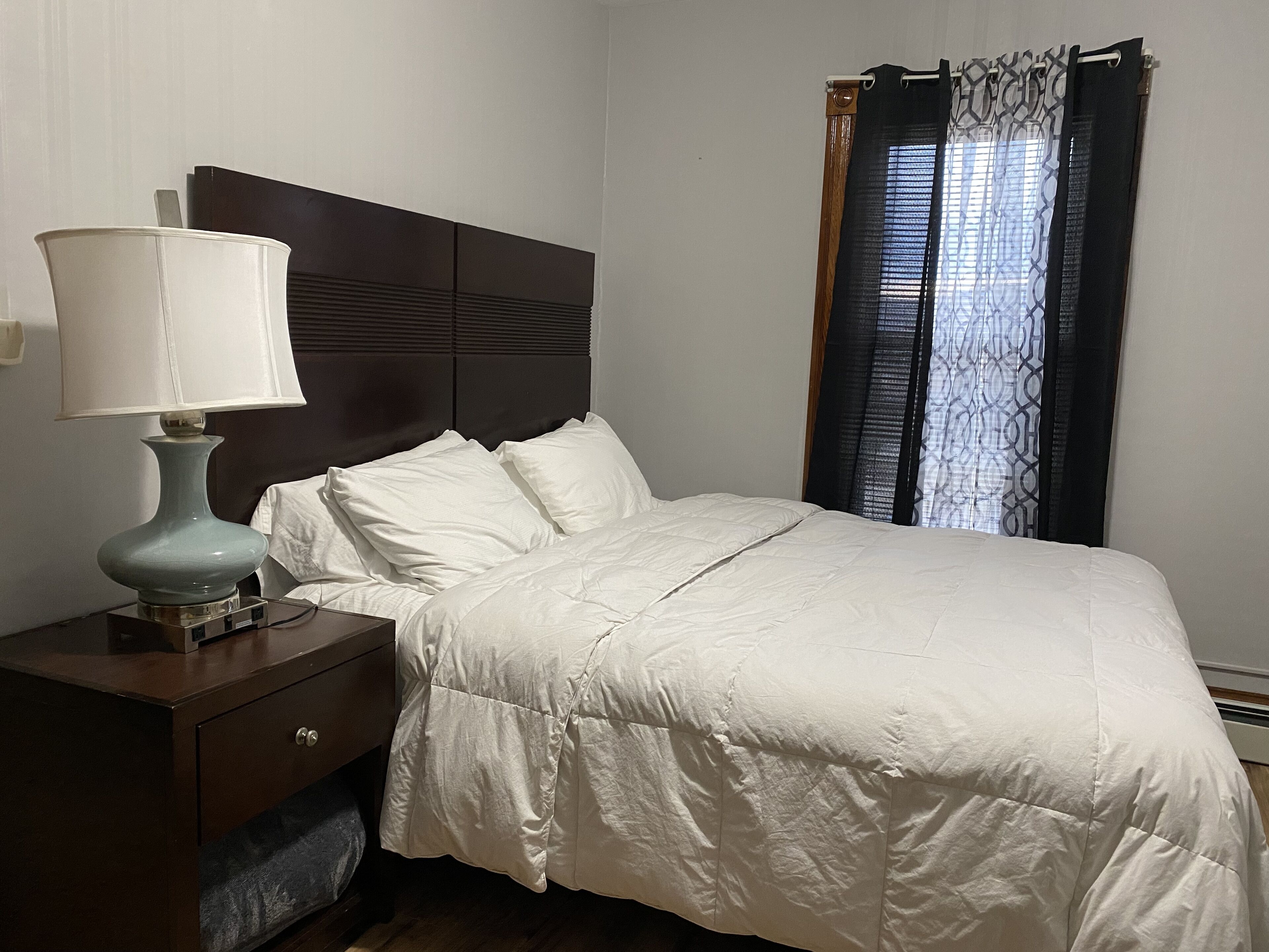 A neatly made bed with white bedding, a bedside table with a lamp, and a window with patterned curtains.