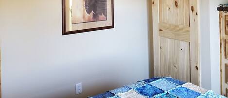 A bedroom with a bed covered in a blue and beige quilt, a ceiling fan, a wooden door, and a framed picture on the wall.