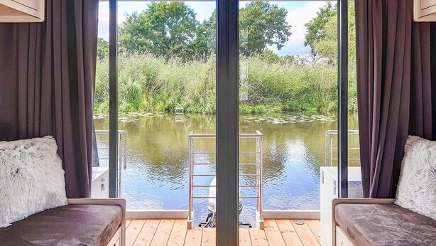 Ein Zimmer mit zwei Sofas, die zu einem großen Fenster mit Blick auf ein Gewässer und eine Holzterrasse zeigen. Eine Metallleiter ist ebenfalls zu sehen.